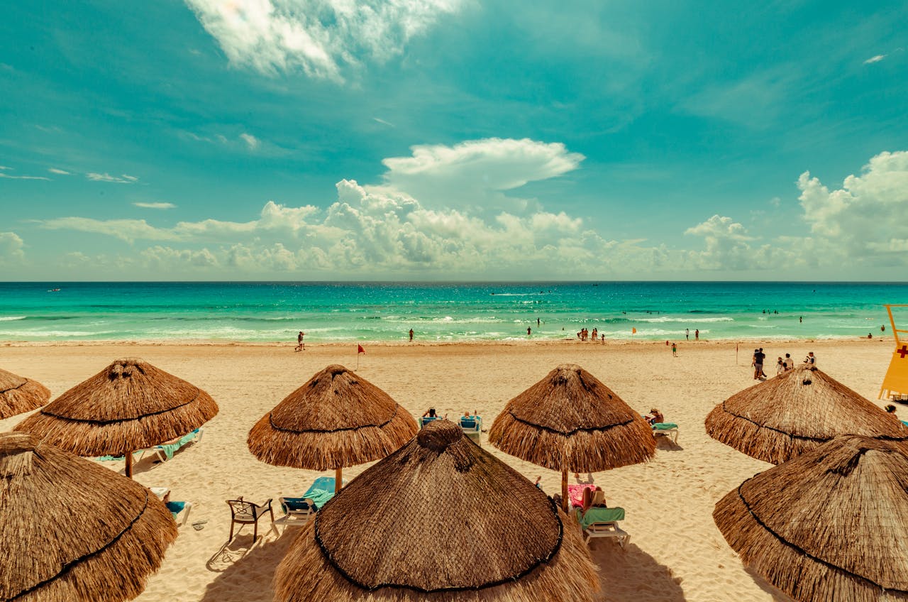 hero-img-01 A vibrant beach scene in Cancun, Mexico with straw umbrellas and turquoise sea under a bright sky.